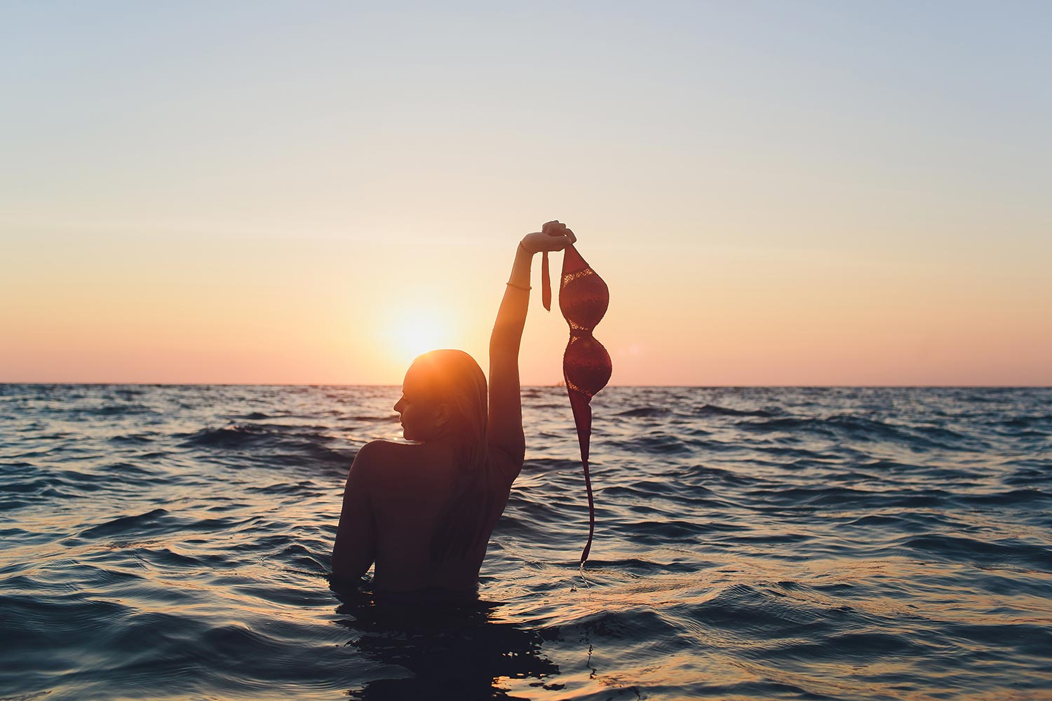 Frau im Wasser zieht sich im Abendrot das Bikinioberteil aus